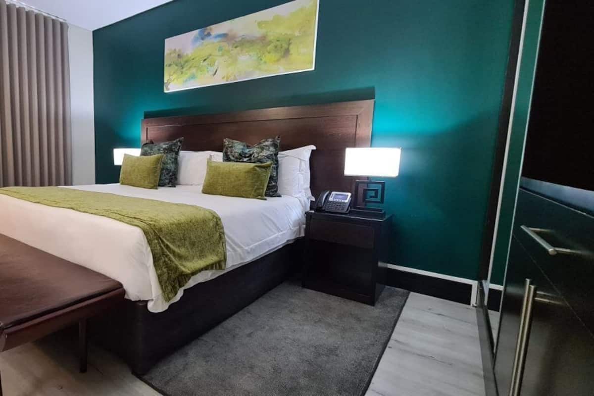 Quarto de hotel com cama, mesas de cabeceira, um banquinho e um quadro na parede.