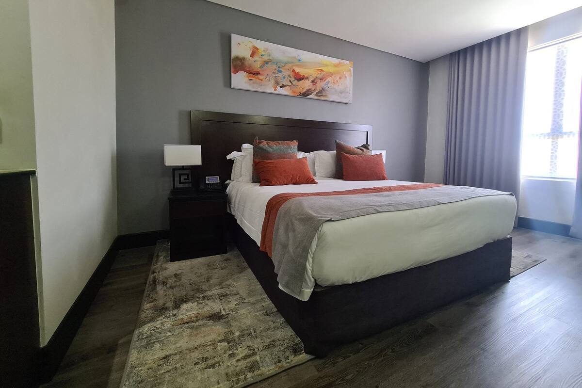 Quarto de hotel com uma cama grande, mesa de cabeceira com abajur, quadro na parede e janela com cortinas.