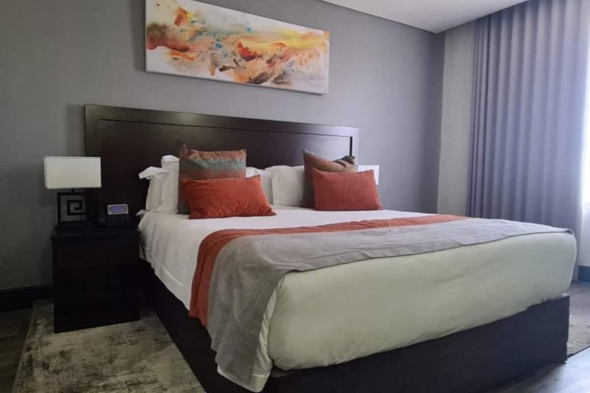 Quarto de hotel moderno com uma cama grande, uma mesa de cabeceira com abajur e um quadro na parede.
