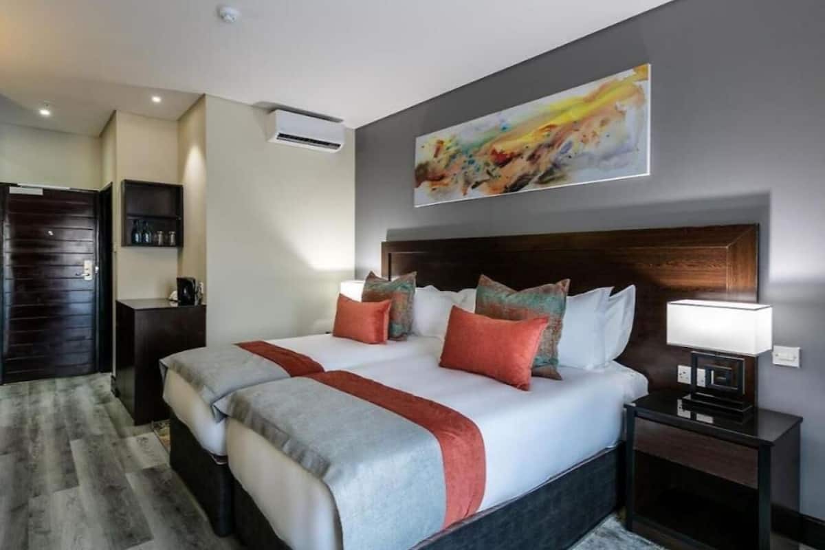 Quarto de hotel com uma cama grande, uma mesa de cabeceira com abajur, ar-condicionado fixado na parede e um quadro sobre a cama.
