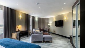 A modern hotel room with a large bed, a seating area with a sofa and ottoman, a desk with a chair, a flat-screen TV mounted on the wall, and a large window with curtains. - Grand Aria Hotel (Gaborone)