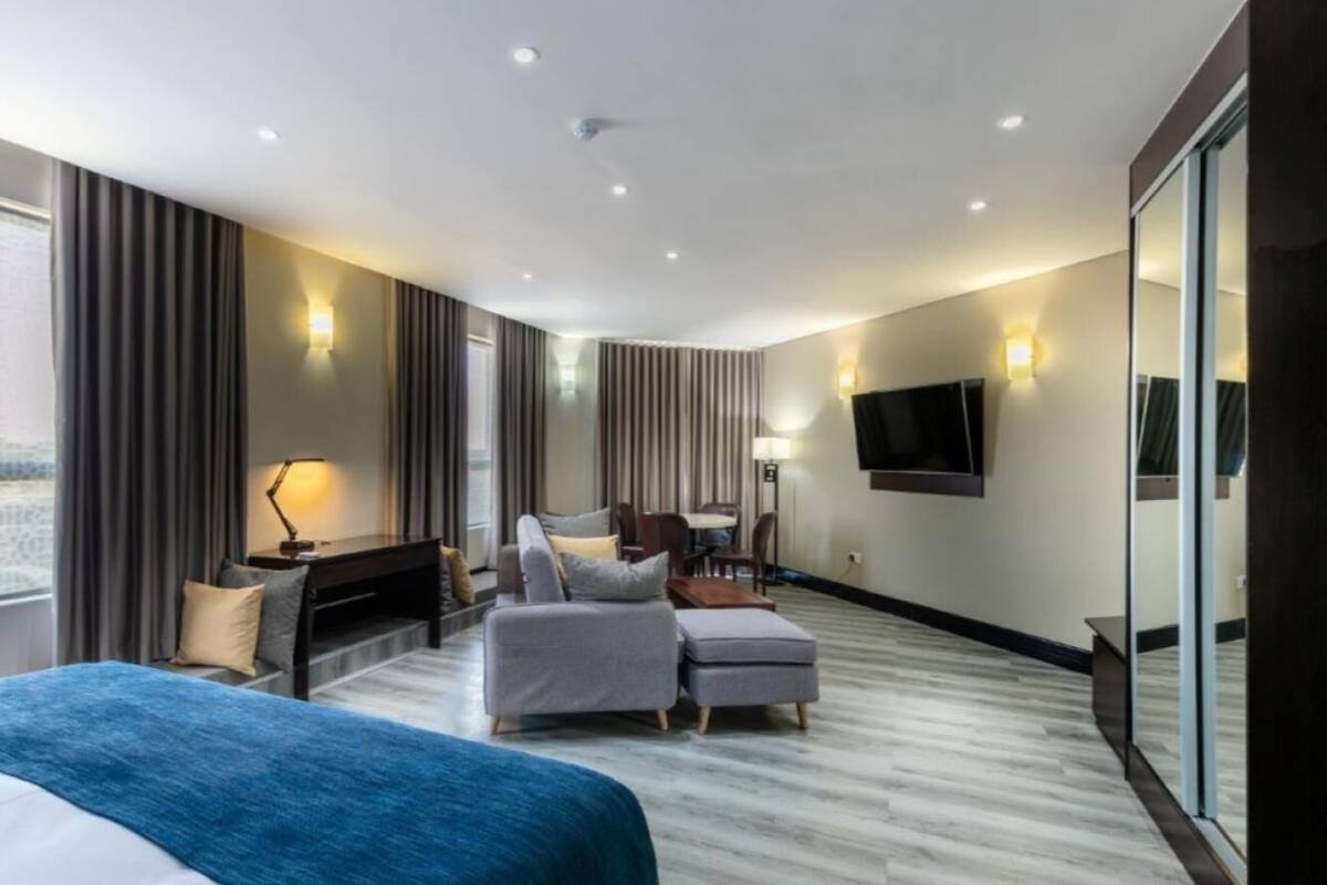 Quarto de hotel moderno com uma cama grande, área de estar com sofá e apoio para os pés, escrivaninha com cadeira, televisão de tela plana na parede e uma ampla janela com cortinas.