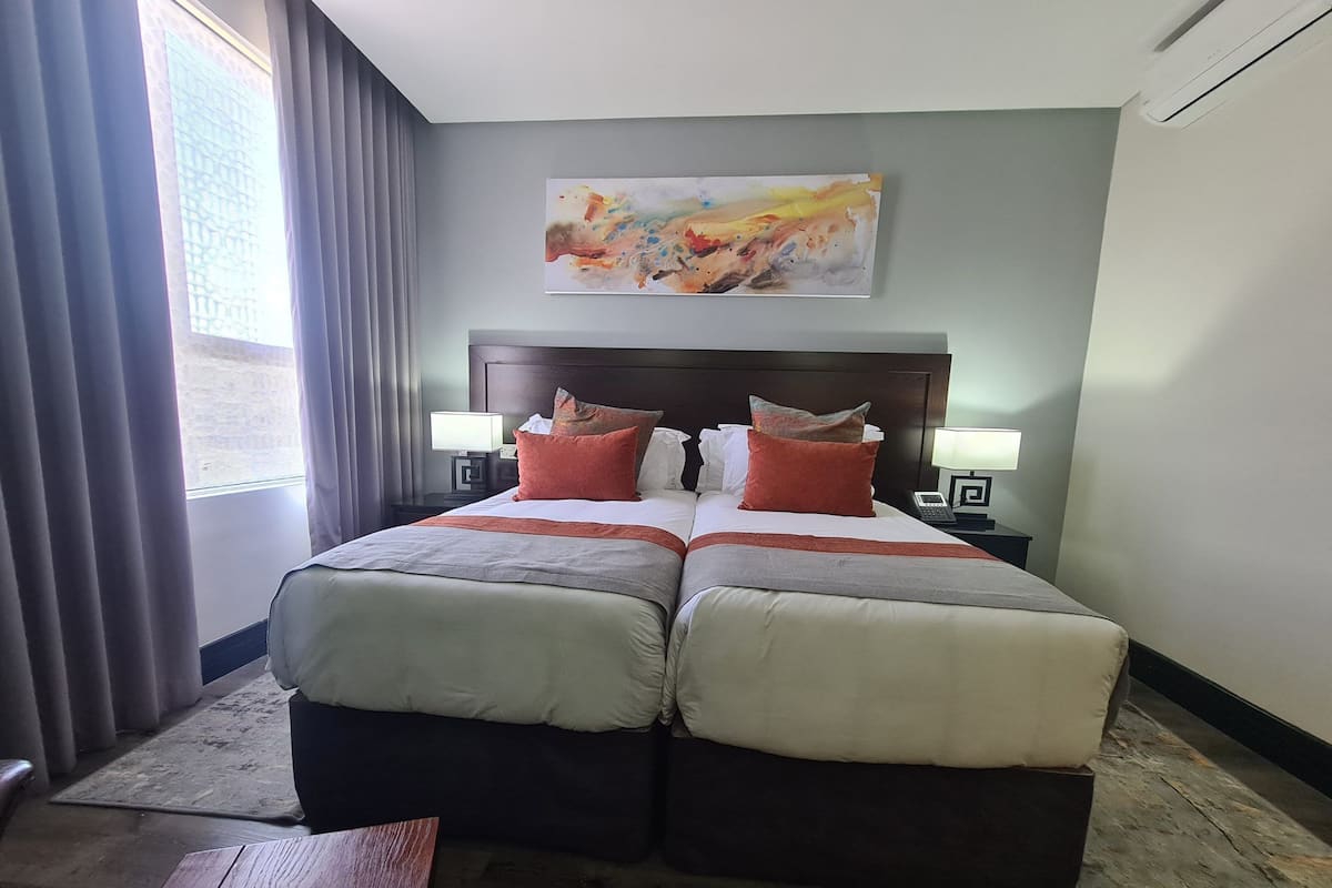 Uma cama de casal com cabeceira de madeira, duas mesas de cabeceira com abajures, um quadro na parede e vista para o exterior através de uma janela com cortinas.