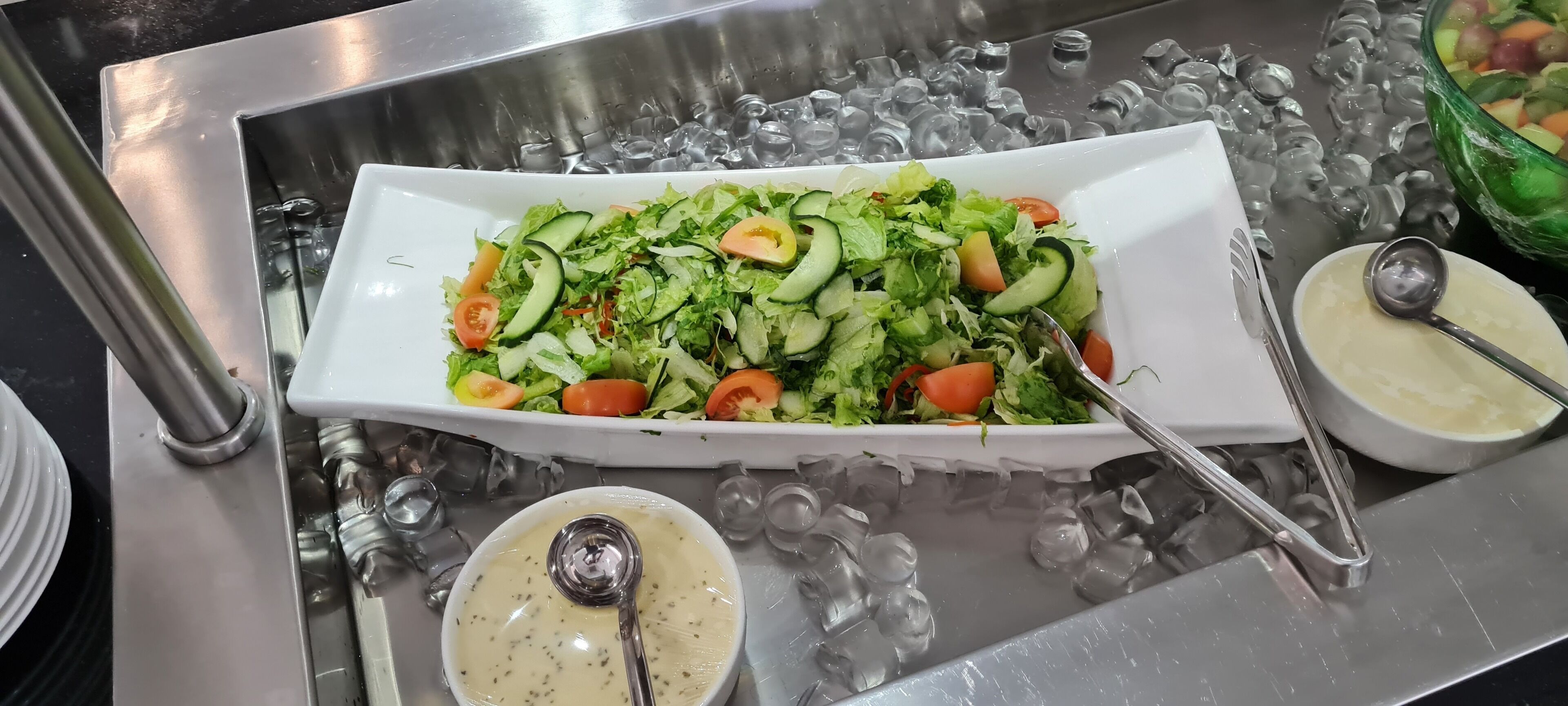 A rectangular white dish filled with a salad of lettuce, cucumber, and tomato slices, accompanied by two small bowls of creamy sauce, all placed on a metallic surface with ice cubes.