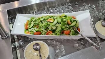 A rectangular white dish filled with a salad of lettuce, cucumber, and tomato slices, accompanied by two small bowls of creamy sauce, all placed on a metallic surface with ice cubes.