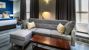 A modern hotel room with a grey sectional sofa, a wooden coffee table, and a bed with a blue accent wall. - Grand Aria Hotel (Gaborone)