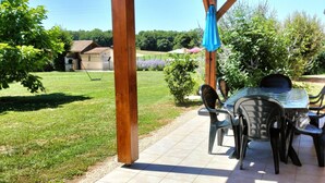 Une terrasse couverte avec une table et des chaises, un barbecue, et une vue sur une zone herbeuse avec des arbres et un bâtiment en arrière-plan.