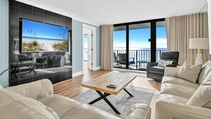 Een ruime woonkamer met een grote televisie, een houten salontafel en een balkon met uitzicht op het strand.