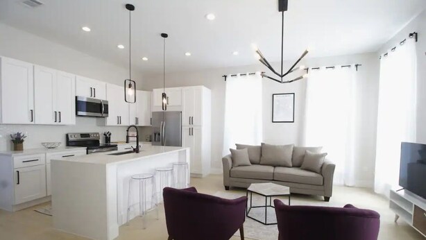 A modern kitchen with white cabinets, a central island, and pendant lighting. There is a living area with a sofa and a TV. - Bright Irish Channel Cottage 10 mins to FQ/Uptown (New Orleans)