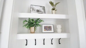 A white wall-mounted shelf with two levels. The top shelf holds a black and white photo, a small potted plant, and a white vase. The bottom shelf has a green plant in a gold pot, a colorful framed photo, and a white ceramic object. - Bright Irish Channel Cottage 10 mins to FQ/Uptown (New Orleans)