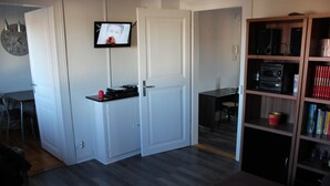 A compact living space with a kitchenette, a television mounted on the wall, and a bookshelf filled with books and decorative items. - APARTMENT GÎTE DE L'OBERHARTH COLMAR ALSACE (Colmar)