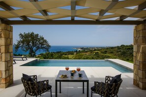 A poolside area with a table set for two, overlooking a scenic view of the sea and greenery.