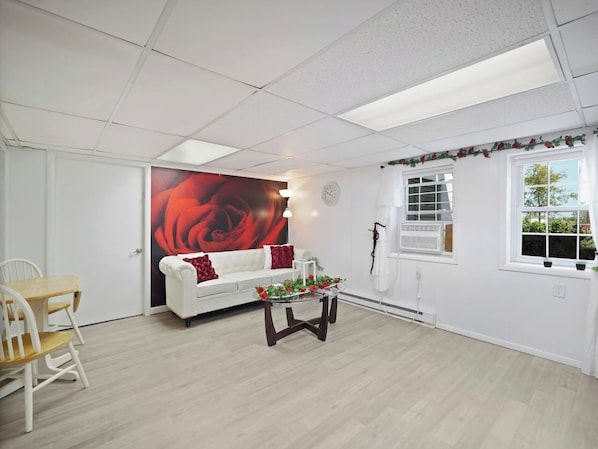 A room with a white sofa, a glass coffee table, a white chair, a large red rose painting, and a window with white curtains.