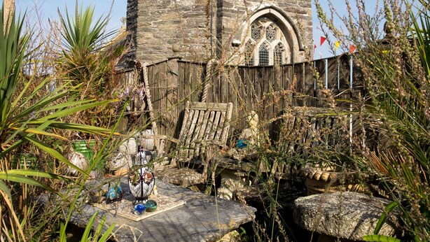 A stone tower with a small arched window, surrounded by overgrown vegetation and a wooden fence. - The Moonbeach Cabin (St Austell)