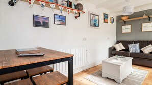 A modern living room with a wooden dining table, a sofa, and a coffee table. There are framed pictures on the wall and a red and white bunting decoration. - The Moonbeach Cabin (St Austell)