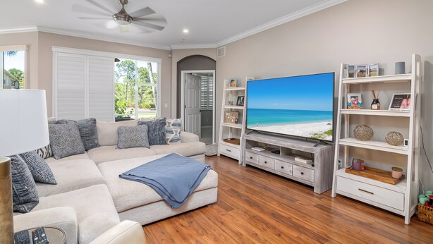 Una sala de estar con un sofá color beige, un televisor de pantalla plana, piso de madera y un ventilador de techo.