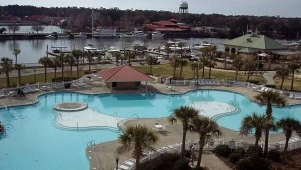 A large swimming pool with a curved design, surrounded by palm trees and a small building with a red roof. - Beautiful 3 Bedroom Condo in Barefoot Landing (North Myrtle Beach)