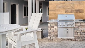 An outdoor patio with a stone barbecue grill and white Adirondack chairs.