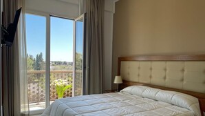 A hotel room with a large bed, a wooden headboard, a nightstand with a lamp, and a sliding glass door leading to a balcony with a view of greenery and buildings. - Hotel Posidonia (Porto Cesareo)