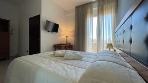 A hotel room with a large bed, a television mounted on the wall, a desk with a chair, and a window with curtains. - Hotel Posidonia (Porto Cesareo)
