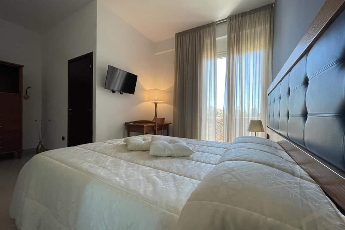 A hotel room with a large bed, a television mounted on the wall, a desk with a chair, and a window with curtains.