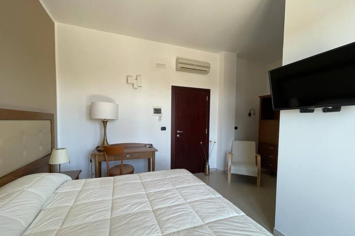 A hotel room with a bed, desk, chair, lamp, TV, and a door.