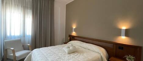 A hotel room with a large bed, a chair, a desk, and two wall-mounted lamps.