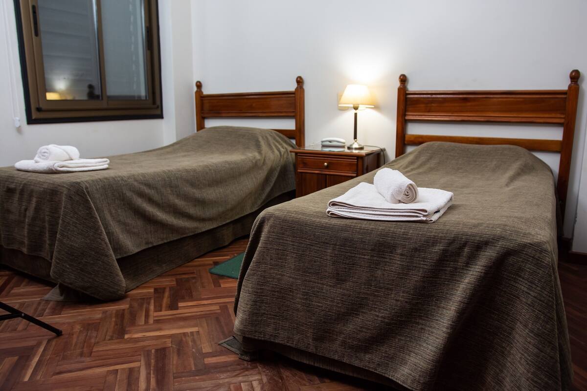A hotel room with two single beds, wooden headboards, a nightstand with a lamp, and a window with blinds.