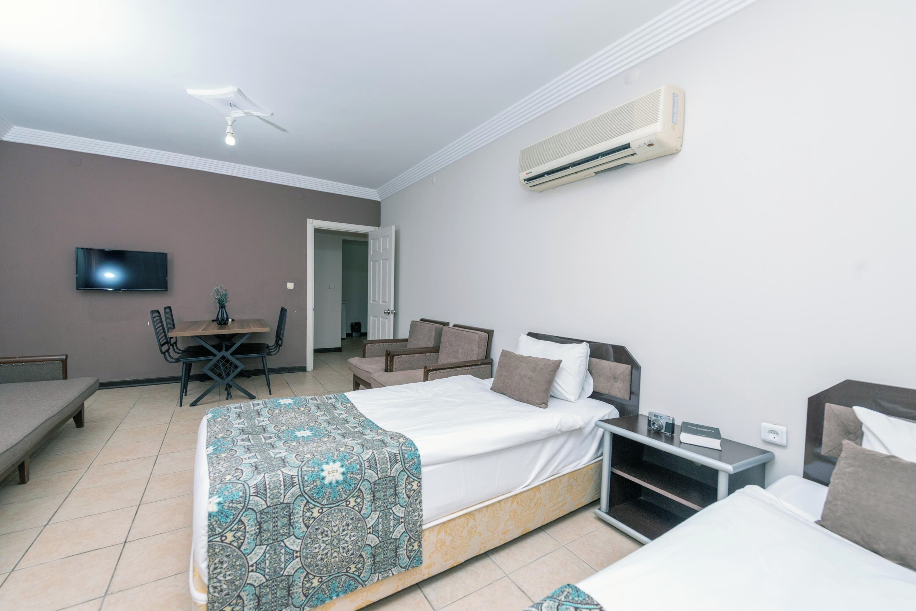 Une chambre d’hôtel moderne équipée d’un lit, d’une table de chevet et d’un bureau avec une chaise. Un téléviseur est fixé au mur et un appareil de climatisation est installé au-dessus du lit.