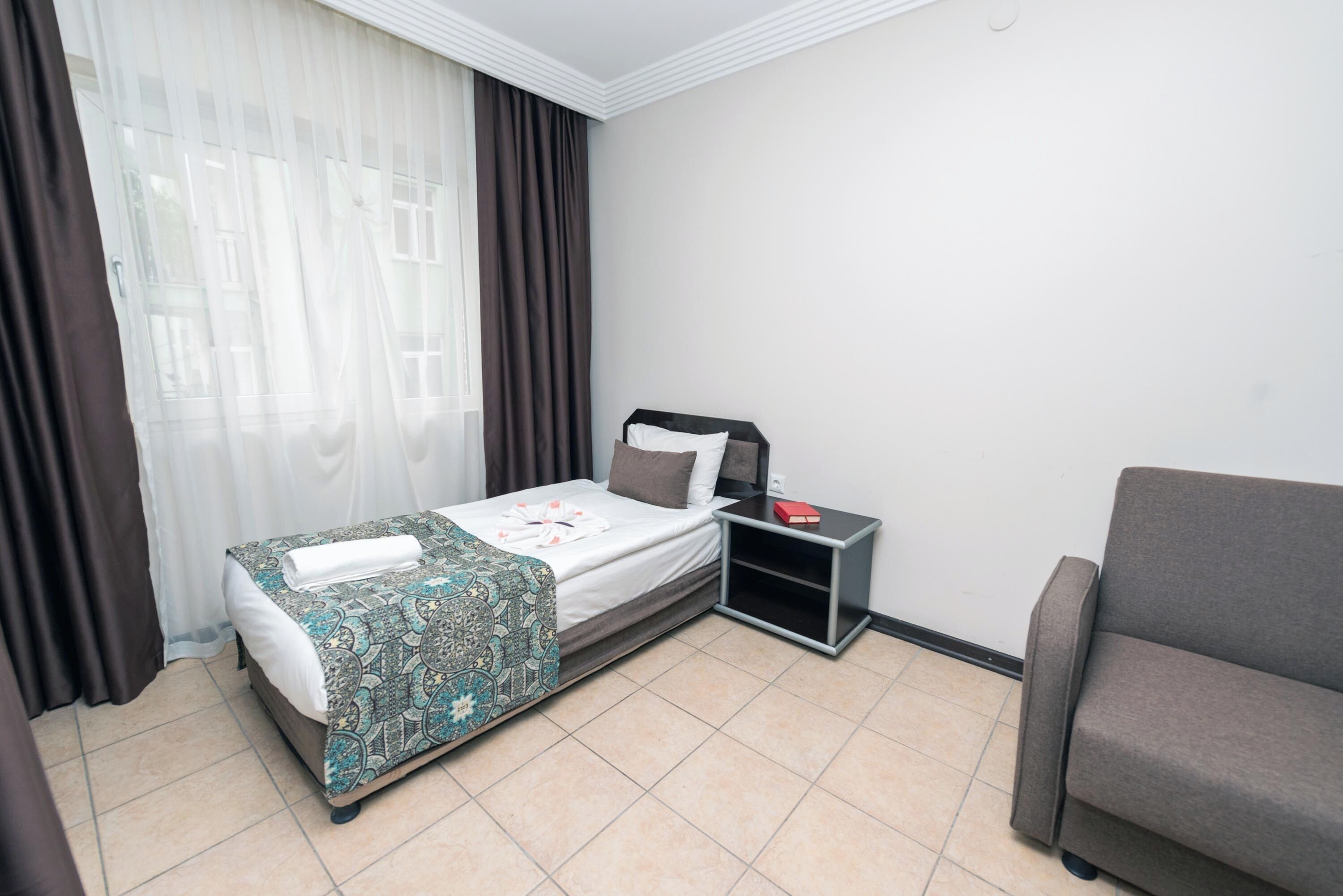 Une chambre d’hôtel avec un lit, une table de chevet, une lampe de lecture, un canapé et une fenêtre avec des rideaux.