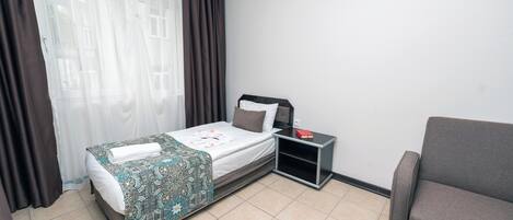 Habitación de hotel con cama, mesita de noche, lámpara de lectura, sofá y ventana con cortinas.