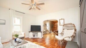 Un salon spacieux doté d’un plancher en bois, d’un ventilateur de plafond, d’une télévision, d’un fauteuil blanc, d’une chaise en osier et d’une petite table.