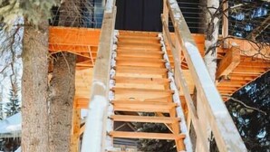 Un escalier en bois mène à une cabane à l’extérieur noir, entourée de neige et d’arbres.