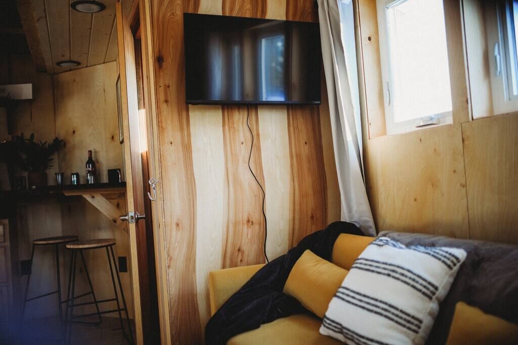 A cozy room with a wooden interior, a flat-screen TV mounted on the wall, a bed with yellow and striped pillows, and a small bar area with stools.