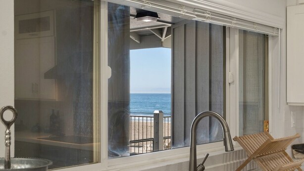 Cozinha com uma grande janela com vista para a praia, pia de metal e uma cadeira de madeira.