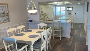 A spacious dining area with a wooden table and white chairs, a kitchen in the background, and a view of the living area. - Luxurious Penthouse condo @ Pointe Santo with magnificent ocean and garden views (Sanibel)