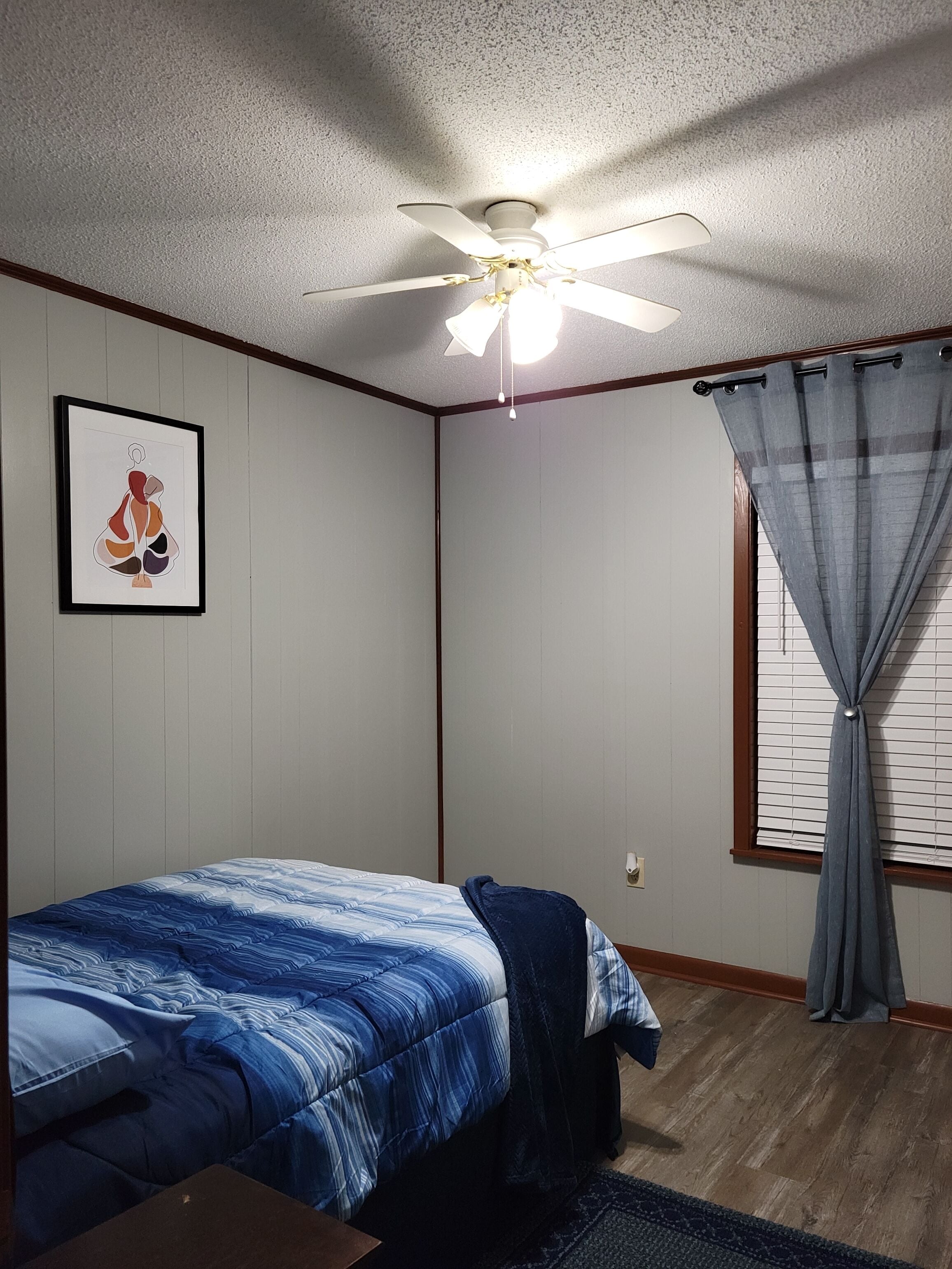 A bedroom with a bed, a ceiling fan, a window with curtains, and a framed picture on the wall.