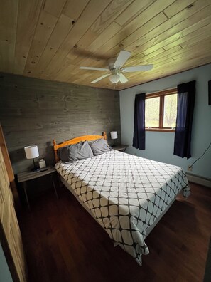A bedroom with a wooden ceiling, a bed with a patterned bedspread, two bedside tables with lamps, a window with curtains, and a wall-mounted outlet. - Secluded Chalet in the Northern Catskills with FIBER Internet and EV hookup (Summit)