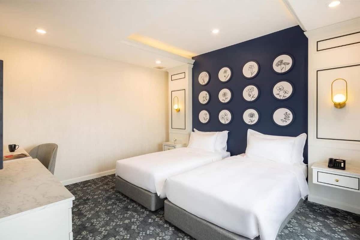 A hotel room with two beds, a desk, and a chair. There are wall-mounted light fixtures and a clock on the nightstand.