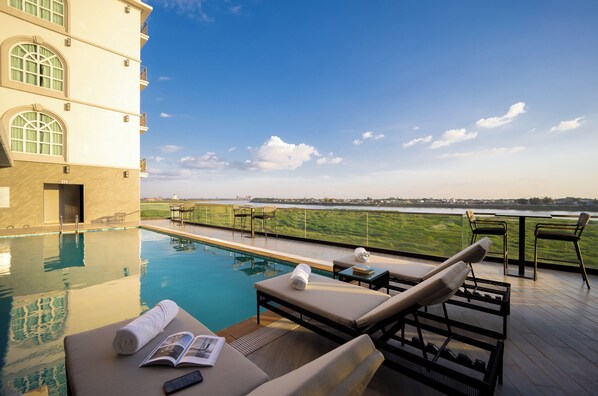 Zona junto a la piscina con sillones, una mesa y un libro, con vistas a un cuerpo de agua y un horizonte urbano distante.