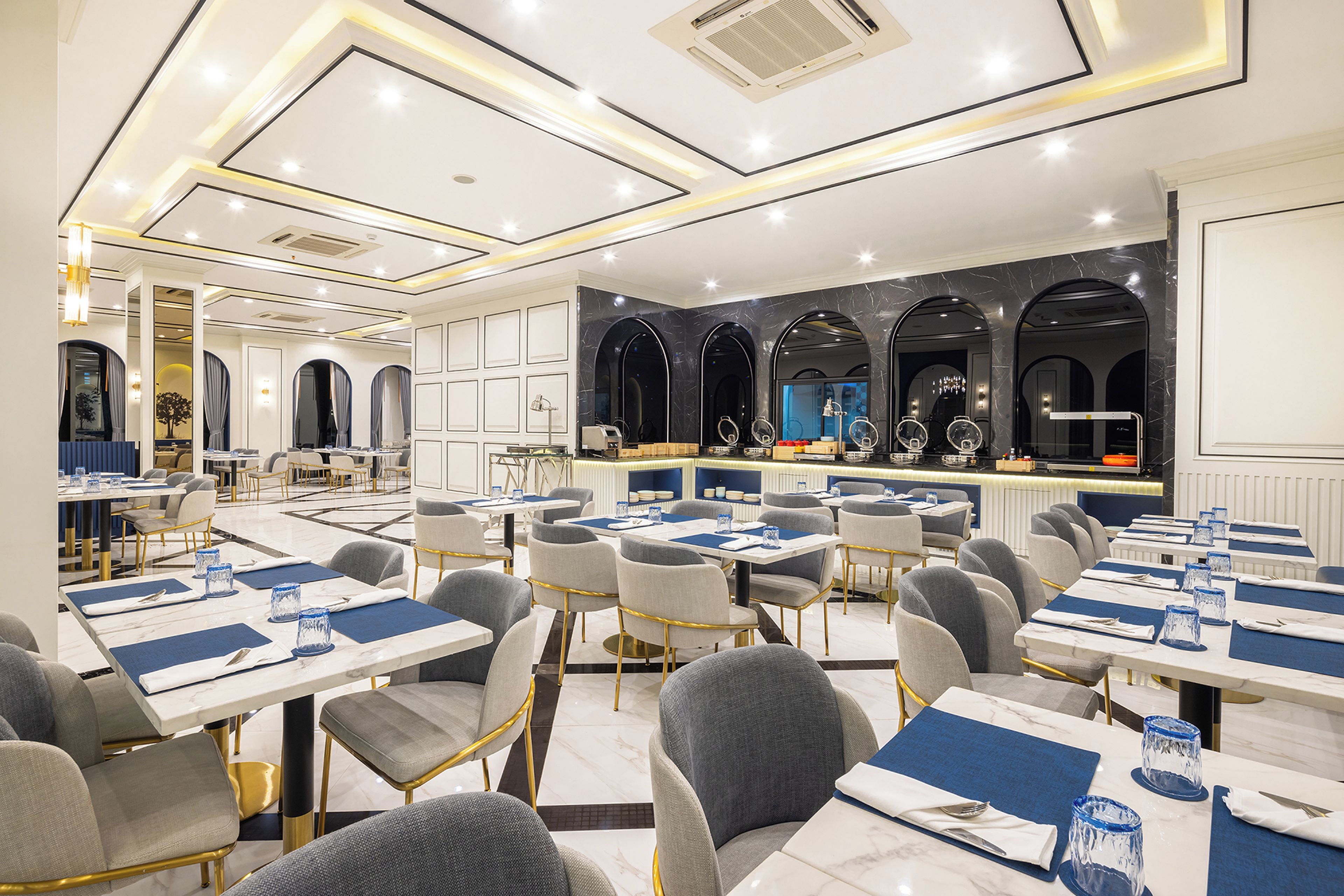 Un comedor moderno con mesas dispuestas para varios comensales, una zona de bar y una cocina al fondo.