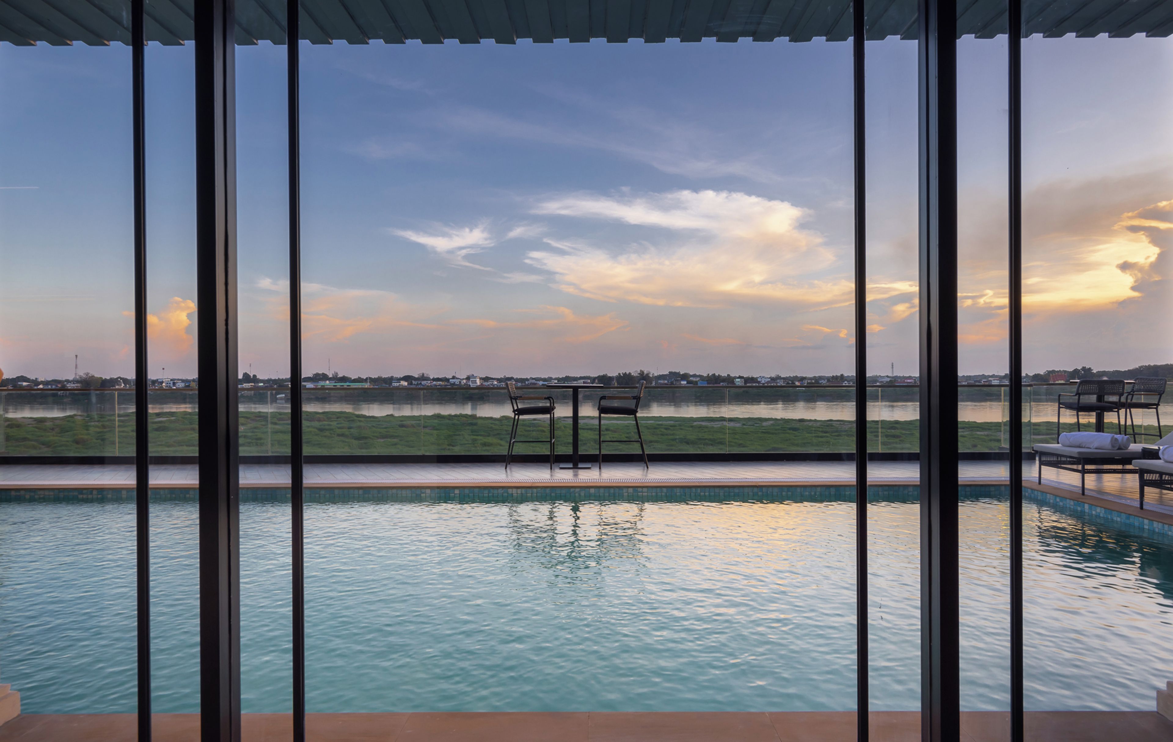 Piscina con dos sillas frente a una vista panorámica de un lago y edificios lejanos al atardecer.