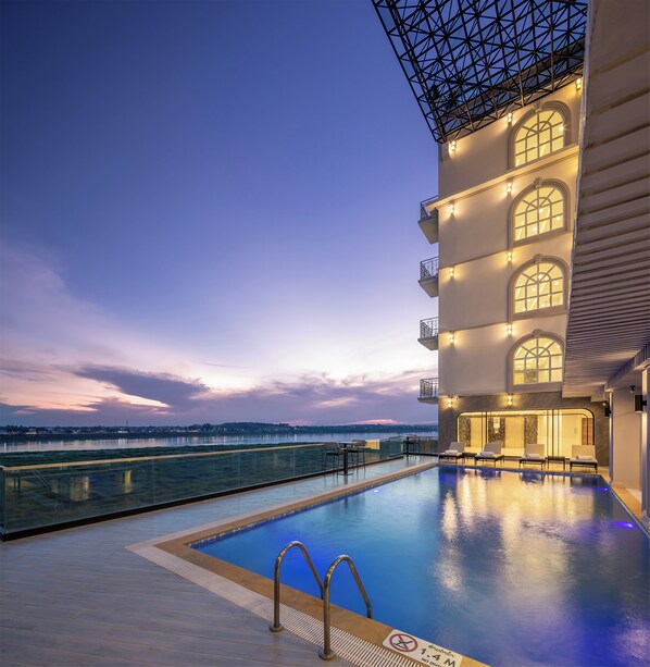 Una piscina en la azotea con barandilla de cristal, vista del horizonte urbano y un edificio con múltiples balcones y grandes ventanales.