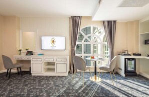 Habitación de hotel con escritorio, dos sillas, una mesita con fruta, televisión y un ventanal grande con cortinas.