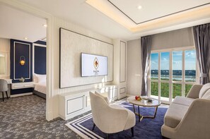 Habitación de hotel con un televisor grande montado en la pared, una zona de estar cómoda con un sofá y sillones, una pequeña mesa redonda con un jarrón de flores y vistas al exterior a través de un amplio ventanal.