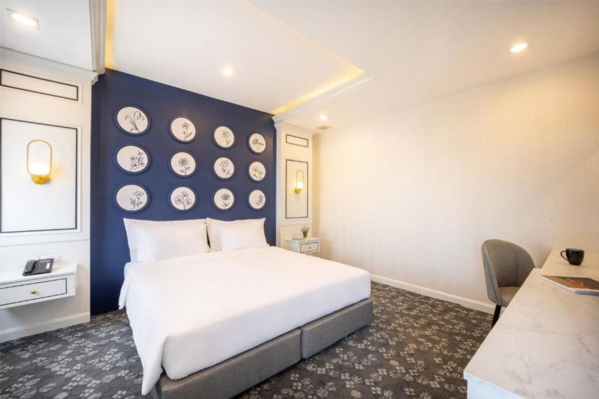 A hotel room with a large bed, a desk, and a chair. There are wall-mounted plates and a small table with a lamp.