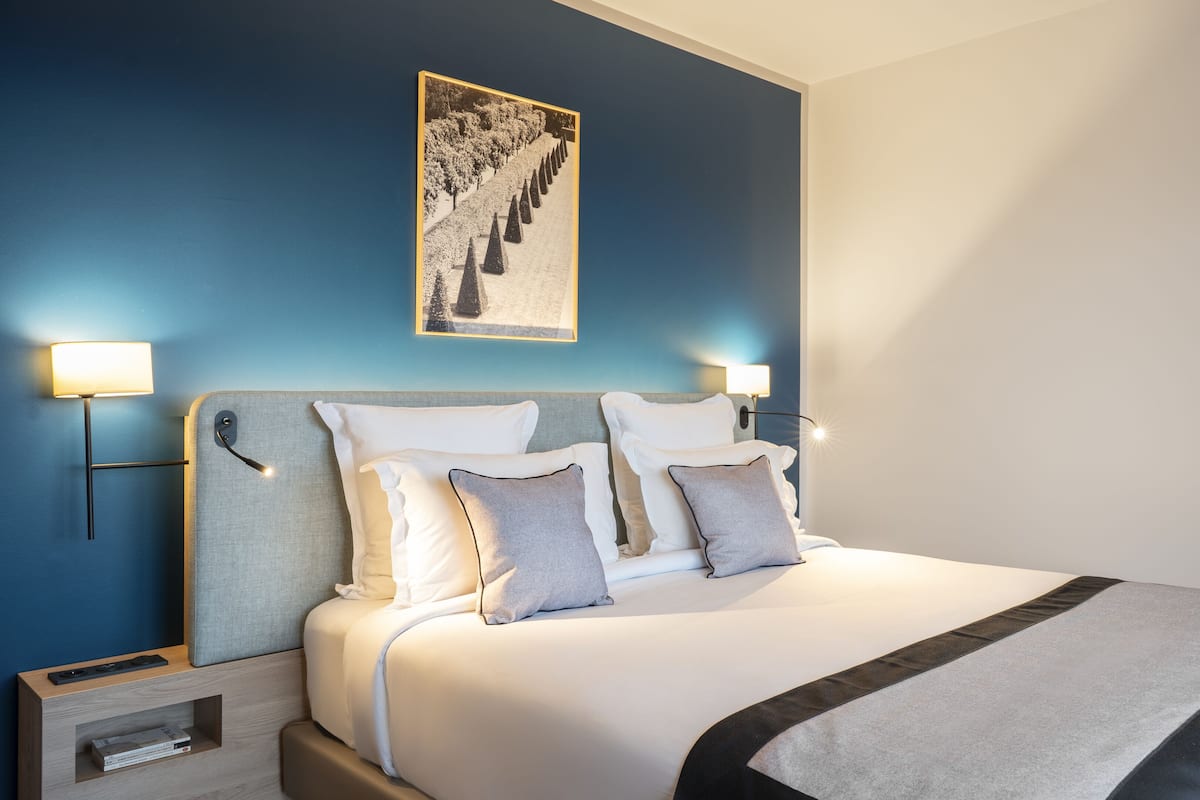 Cama bem arrumada com roupa de cama branca e cinza, duas luminárias de cabeceira, um quadro na parede e uma pequena mesa de cabeceira de madeira com uma luminária.