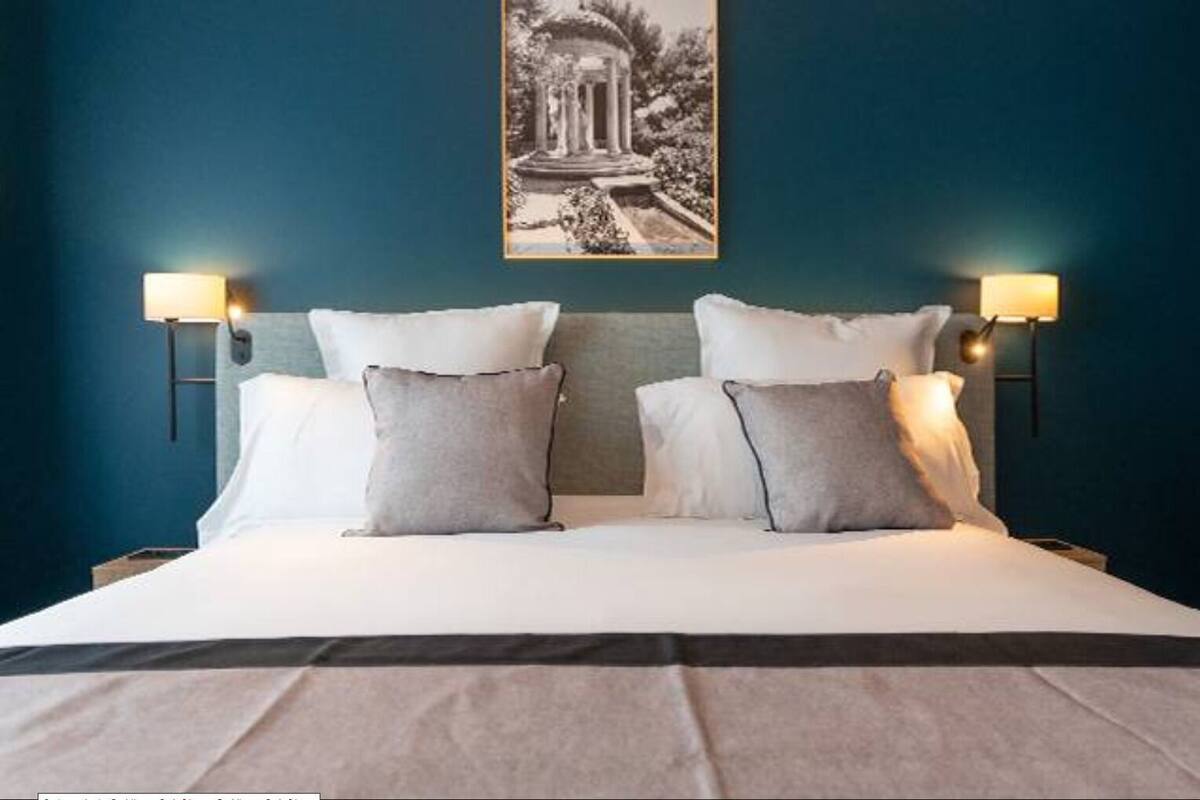Uma cama com roupa de cama branca e duas almofadas cinzas, com luminárias fixadas na parede, em um quarto com paredes verde-água e um quadro emoldurado acima da cama.