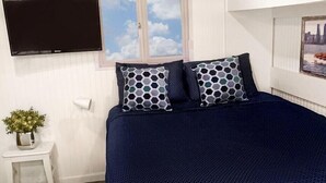 A bedroom with a large bed, a television mounted on the wall, a small bedside table with a plant, and a window with a view of the sky. - Miramar Breeze (Miramar)
