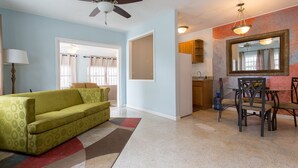 Un salon avec un canapé vert, un ventilateur de plafond, un coin repas avec une table et des chaises, et une cuisine visible à travers une porte ouverte.